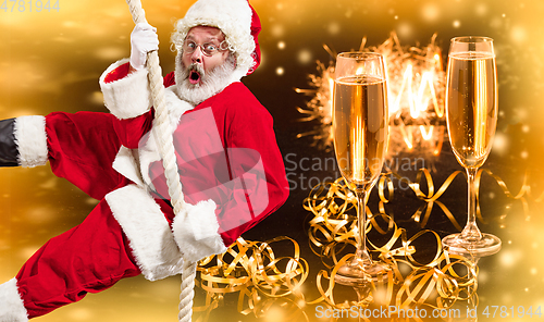 Image of Happy Christmas Santa Claus climbing on rope with glasses of champagne in background