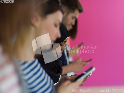 Image of diverse teenagers use mobile devices while posing for a studio photo in front of a pink background