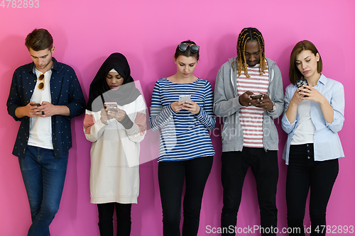 Image of diverse teenagers use mobile devices while posing for a studio photo in front of a pink background