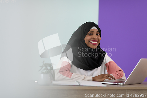 Image of afro muslim woman wearing a hijab sits smiling in her home office and uses a laptop