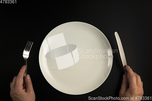 Image of Hands with fork and knife and white plate on black background in flat lay