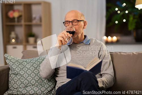 Image of senior man with book drinking red wine at home