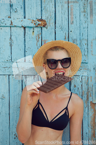 Image of Young attractive woman in sunglasses and in straw hat eating chocolate on a blue background. Film efect