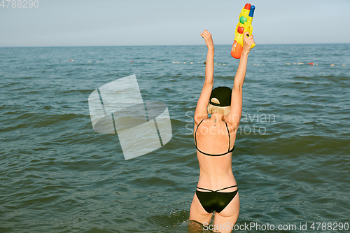 Image of Happy young woman in a cap with the word queen playing with water gun. Film effect