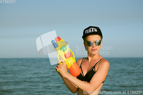 Image of Happy young woman in a cap with the word queen playing with water gun. Film effect