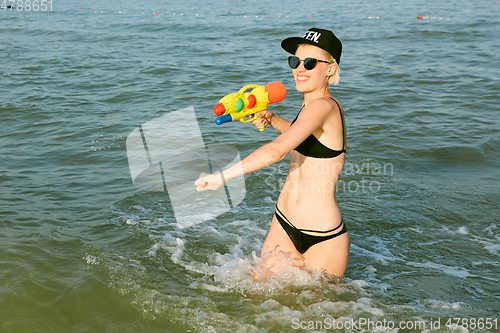 Image of Happy young woman in a cap with the word queen playing with water gun. Film effect