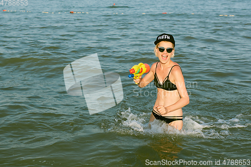 Image of Happy young woman in a cap with the word queen playing with water gun. Film effect