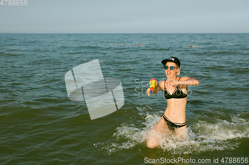 Image of Happy young woman in a cap with the word queen playing with water gun. Film effect