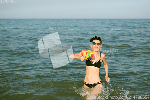 Image of Happy young woman in a cap with the word queen playing with water gun. Film effect