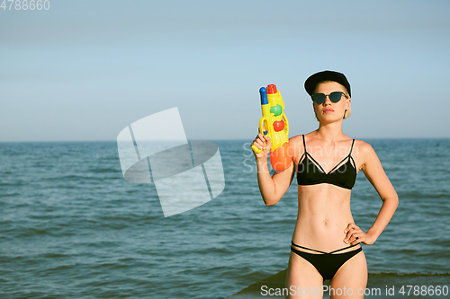 Image of Happy young woman in a cap with the word queen playing with water gun. Film effect