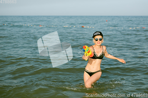 Image of Happy young woman in a cap with the word queen playing with water gun. Film effect