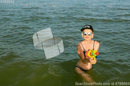 Image of Happy young woman in a cap with the word queen playing with water gun. Film effect