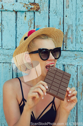 Image of Young attractive woman in sunglasses and in straw hat eating chocolate on a blue background. Film efect