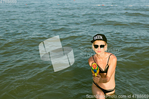 Image of Happy young woman in a cap with the word queen playing with water gun. Film effect