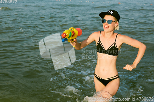 Image of Happy young woman in a cap with the word queen playing with water gun. Film effect