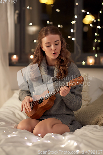 Image of happy young woman playing guitar in bed at home