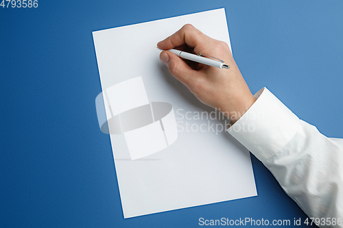 Image of Male hand holding pen and writing on empty sheet on blue background for text or design