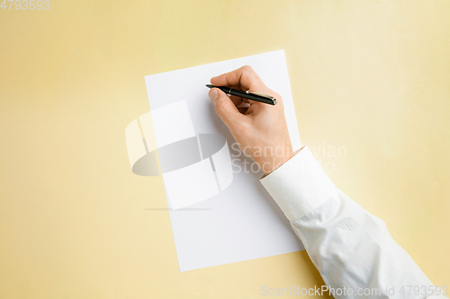 Image of Male hand holding pen and writing on empty sheet on yellow background for text or design