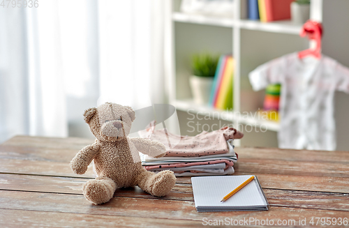 Image of baby clothes, teddy bear toy and notebook