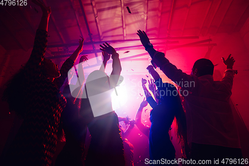 Image of A crowd of people in silhouette raises their hands against colorful neon light on party background