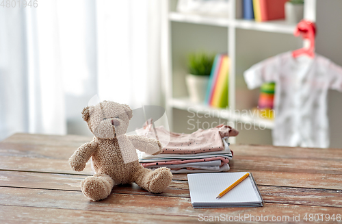 Image of baby clothes, teddy bear toy and notebook