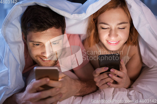 Image of happy couple using smartphones in bed at night