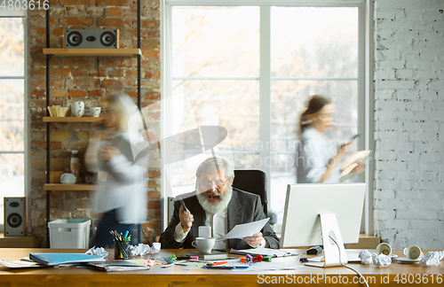 Image of Nervous and tired boss at his workplace busy while people moving near blurred