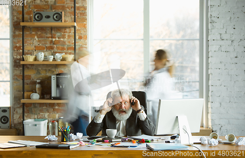 Image of Nervous and tired boss at his workplace busy while people moving near blurred