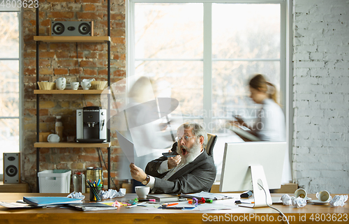 Image of Nervous and tired boss at his workplace busy while people moving near blurred