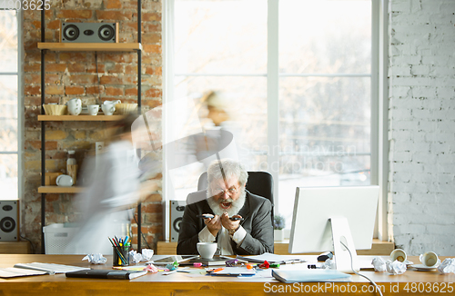 Image of Nervous and tired boss at his workplace busy while people moving near blurred