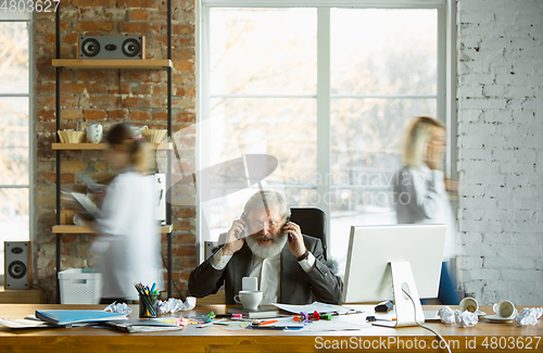 Image of Nervous and tired boss at his workplace busy while people moving near blurred