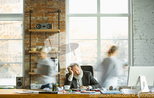 Image of Nervous and tired boss at his workplace busy while people moving near blurred