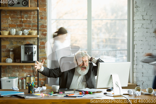 Image of Nervous and tired boss at his workplace busy while people moving near blurred