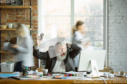 Image of Nervous and tired boss at his workplace busy while people moving near blurred