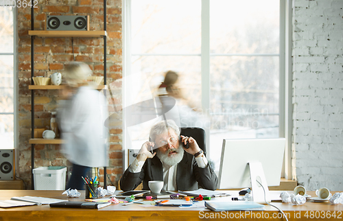 Image of Nervous and tired boss at his workplace busy while people moving near blurred