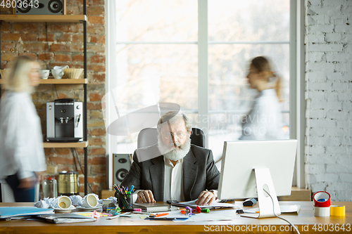 Image of Nervous and tired boss at his workplace busy while people moving near blurred