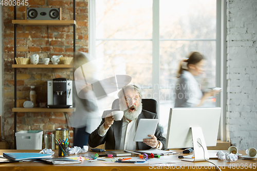 Image of Nervous and tired boss at his workplace busy while people moving near blurred