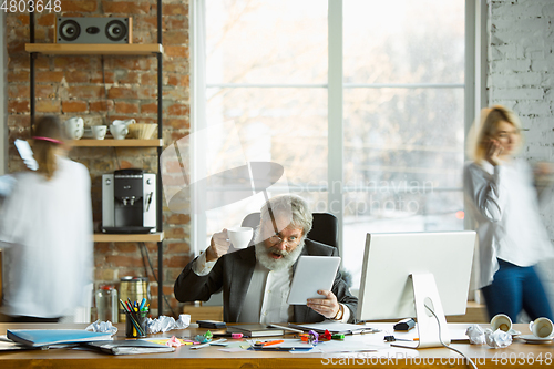 Image of Nervous and tired boss at his workplace busy while people moving near blurred