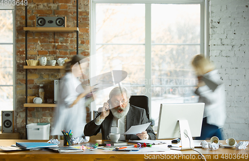 Image of Nervous and tired boss at his workplace busy while people moving near blurred