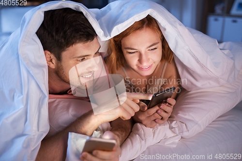 Image of happy couple using smartphones in bed at night