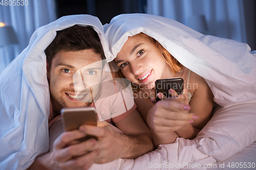 Image of happy couple using smartphones in bed at night