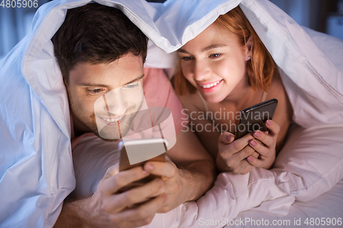 Image of happy couple using smartphones in bed at night