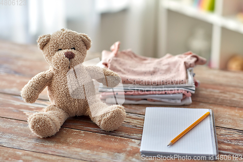 Image of baby clothes, teddy bear toy and notebook