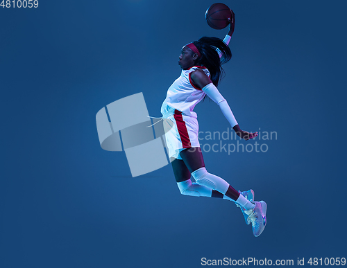 Image of Beautiful african-american female basketball player in motion and action in neon light on blue background. Concept of healthy lifestyle, professional sport, hobby.