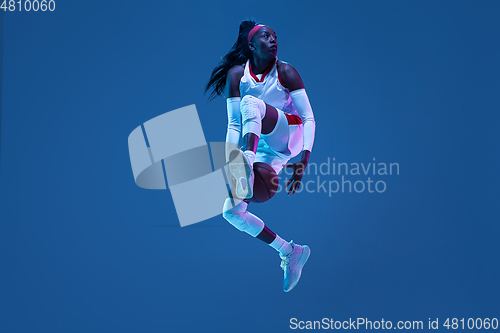 Image of Beautiful african-american female basketball player in motion and action in neon light on blue background. Concept of healthy lifestyle, professional sport, hobby.