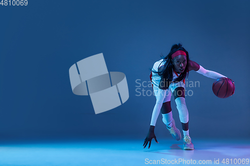 Image of Beautiful african-american female basketball player in motion and action in neon light on blue background. Concept of healthy lifestyle, professional sport, hobby.