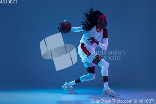 Image of Beautiful african-american female basketball player in motion and action in neon light on blue background. Concept of healthy lifestyle, professional sport, hobby.