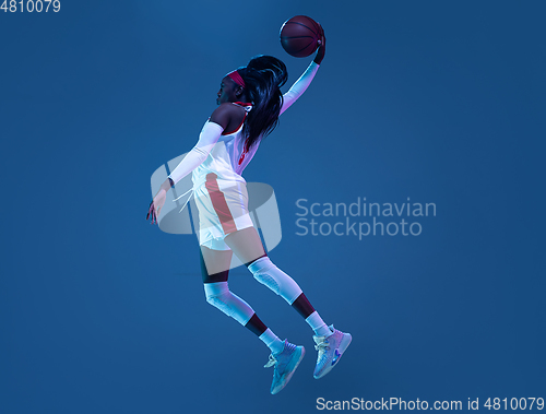 Image of Beautiful african-american female basketball player in motion and action in neon light on blue background. Concept of healthy lifestyle, professional sport, hobby.