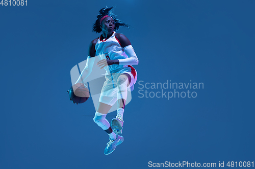 Image of Beautiful african-american female basketball player in motion and action in neon light on blue background. Concept of healthy lifestyle, professional sport, hobby.