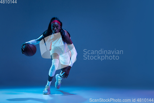 Image of Beautiful african-american female basketball player in motion and action in neon light on blue background. Concept of healthy lifestyle, professional sport, hobby.
